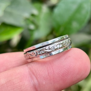 May include: A silver spinner ring with an ornate floral design. The ring has three bands, with the center band featuring the detailed pattern. The ring is held up by a finger, with a blurred green background.