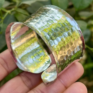 Peut inclure: Un large bracelet manchette argenté avec une texture martelée. Le bracelet a une forme incurvée et un design ouvert, permettant de l'enfiler et de l'enlever facilement. La surface reflète la verdure environnante.