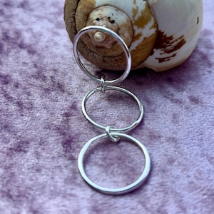 May include: A silver ring set comprising three linked circular rings. The rings are joined by small, delicate knots. The rings are presented on a textured, purple surface, with a seashell in the background.