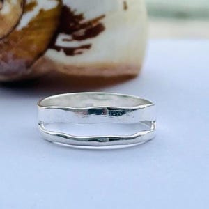 Op de afbeelding: Een zilveren ring met een uniek ontwerp met dubbele band. De ring heeft een getextureerde, gehamerde uitstraling, waardoor hij een handgemaakte uitstraling heeft. De banden zijn gescheiden, waardoor een open ruimte in het midden ontstaat. De ring staat tegen een zachte, vage achtergrond.