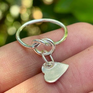 Puede incluir: Un anillo de plata con un diseño de nudo y un pequeño dije de corazón. El anillo está hecho de metal brillante y tiene una apariencia delicada. El dije de corazón cuelga del nudo, agregando un toque de sentimentalismo a la joya.