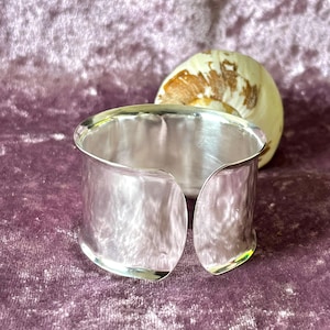 May include: A wide, open silver cuff bracelet with a textured surface. The bracelet is displayed on a purple velvet surface, with a seashell in the background. The cuff is approximately 2 inches wide.