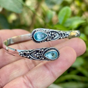 May include: A silver-toned cuff bracelet with two teardrop-shaped, light blue gemstones. The bracelet features ornate detailing around the gemstones and along the band. The gemstones are set in a decorative, openwork setting. The bracelet is shown against a blurred green background.