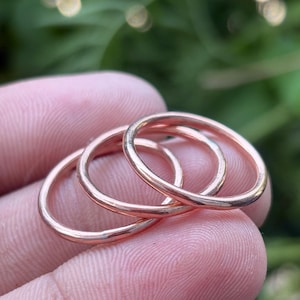 Puede incluir: Tres delicados anillos de oro rosa están entrelazados, mostrando un diseño minimalista. Los anillos se sostienen entre los dedos, con un fondo verde borroso. Los anillos parecen tener aproximadamente 0,25 cm de ancho.