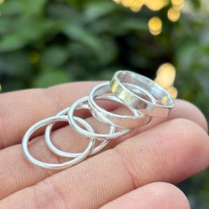 May include: A collection of silver rings, varying in thickness, are displayed. The rings are stacked and slightly angled, showcasing their simple, minimalist design. The rings are held in a hand, with a blurred green background.