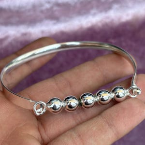 Könnte beinhalten: Ein silbernes Armreif-Armband mit sechs runden, polierten Perlen. Das Armband hat ein schlichtes, elegantes Design und wird in einer Hand vor einem lila Hintergrund gehalten. Die Perlen sind gleichmäßig über das Armband verteilt.