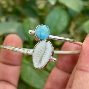 Pode incluir: Uma pulseira prateada com uma pedra preciosa azul clara e uma concha branca. A pulseira tem um design simples e elegante com um acabamento polido. As pedras preciosas e a concha são os pontos focais da pulseira.