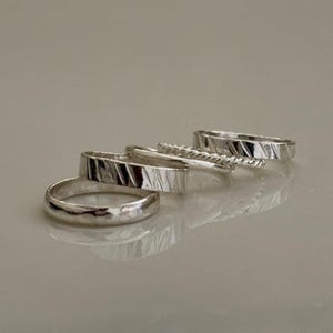 May include: A collection of silver rings in various designs. The rings include a smooth band, a textured band, a twisted rope design, and a band with a diagonal etched pattern. The rings are displayed on a light gray surface.