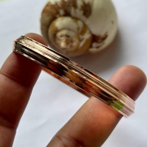 May include: A rose gold bangle bracelet with a layered design. The bracelet is held in a hand, with a seashell in the background. The bangle has a polished finish and a simple, elegant style.