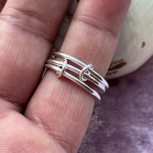 May include: A silver ring with a unique design. The ring features three parallel bands connected by two knot-like elements. The ring is worn on a finger, showcasing its intricate details and polished finish. The jewelry is a simple yet elegant accessory.