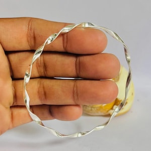 May include: A silver-colored bangle bracelet with a twisted design. The bracelet is circular and appears to be made of a shiny metal. The bangle is held in a person's hand, with a blurred background.