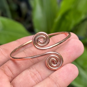 Op de afbeelding: Een gepolijste koperkleurige metalen armband met een open ontwerp en twee symmetrische, spiraalvormige uiteinden. De armband is gemaakt van een gladde, afgeronde draad die buigt om de cirkelvormige spiraalmotieven aan de uiteinden te vormen.