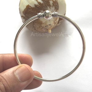 Op de afbeelding: Een zilveren armband met een centrale bolvormige kraal en decoratieve accenten. De armband wordt in een hand gehouden, met de tekst "ArtisanjewelArt" zichtbaar. Een eenvoudig, elegant ontwerp.