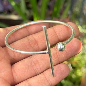 May include: A silver-colored bracelet with a minimalist design. The bracelet features a curved band with a vertical bar and a small ball accent. The jewelry is held in a hand, with a blurred green background.
