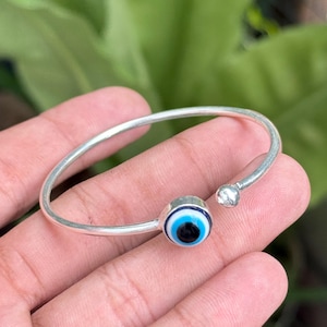 Könnte beinhalten: Ein silberfarbenes, offenes Armband mit einem kreisförmigen Nazar-Amulett an einem Ende und einer kleinen silberfarbenen Kugel am anderen Ende. Das Amulett zeigt konzentrische Ringe in Dunkelblau, Hellblau und Weiß auf einer runden Basis.