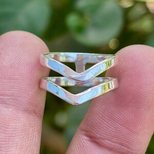 Pode incluir: Dois anéis prateados com um design em forma de V são segurados entre os dedos. Os anéis têm uma superfície polida e reflexiva. O fundo é verde desfocado.