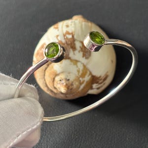 May include: A silver cuff bracelet with two round, bright green peridot gemstones. The bracelet is displayed against a seashell on a dark surface. The gemstones are set in silver bezels.