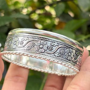 Puede incluir: Un brazalete plateado con un diseño floral. El brazalete presenta tallas detalladas de flores y hojas alrededor de la banda. Los bordes tienen un diseño de cuerda trenzada. El fondo es verde borroso.