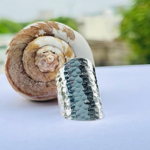 Op de afbeelding: Een zilverkleurige ring met een gehamerde textuur. De ring staat naast een grote, spiraalvormige zeeschelp met bruine en witte markeringen. De ring heeft een brede band en een licht gebogen vorm.