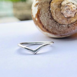 Könnte beinhalten: Ein silberfarbener Ring mit V-förmigem Design. Der Ring ist schlicht und elegant, mit glatter, polierter Oberfläche. Der Ring befindet sich vor einem weißen Hintergrund, mit einer Muschel im Hintergrund.