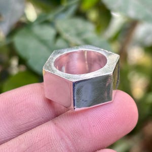 Peut inclure: Une bague hexagonale argentée avec une finition polie. La bague a un centre creux et est tenue par le doigt d'une personne. L'arrière-plan est un feuillage vert flou.
