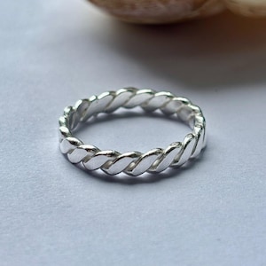 May include: A silver ring featuring a twisted rope pattern. The ring is circular and rests on a pale grey surface. The design consists of repeating, interwoven segments, giving it a textured look. The ring is approximately 0.2 cm wide.