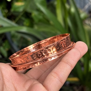 Może przedstawiać: Miedziana bransoletka typu bangle o młotkowanej fakturze z serią kolistych wgłębień na powierzchni. Metal ma polerowane, odblaskowe wykończenie z wyraźnym, wypukłym żebrem biegnącym poziomo wokół środka.