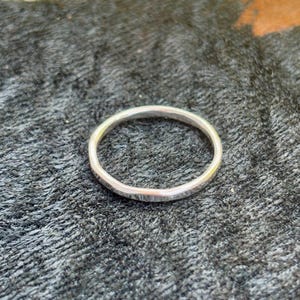 May include: A silver ring with a textured surface. The ring is circular and appears to be made of metal. The ring is set against a dark, textured background.