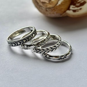 Könnte beinhalten: Vier silberne Ringe mit detaillierten Mustern liegen auf einer weißen Oberfläche. Die Ringe weisen verschiedene Designs auf, darunter Blumen- und Blättermotive. Die Ringe sind wahrscheinlich aus Sterlingsilber und für den täglichen Gebrauch geeignet.