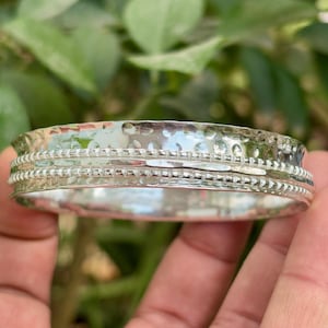 Könnte beinhalten: Ein silbernes Armreif-Armband mit gehämmerten Texturen und Perlendetails. Das Armband hat eine glänzende, reflektierende Oberfläche und ist für das Handgelenk konzipiert. Das Design ist schlicht und elegant, geeignet für den Alltag.