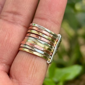May include: A multi-tone ring with a silver base and multiple bands in gold, rose gold, and silver. The ring is worn on a finger, showcasing the different metal colours and textures. The bands are stacked, creating a unique and eye-catching design.