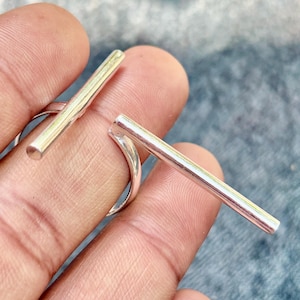 Può includere: Un anello color argento dal design minimalista. L'anello presenta una barra dritta sulla parte superiore, con un'interruzione al centro. L'anello è mostrato su un dito.