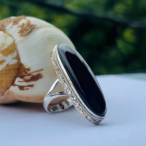 May include: A silver ring with an oval, polished black stone. The ring has a detailed, patterned border around the stone. The band is a simple, polished silver. The ring is set against a blurred background.