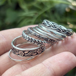 Può includere: Una collezione di anelli d'argento con vari design, tra cui fasce floreali, intrecciate e decorate. Gli anelli sono impilati, mostrando i loro dettagli e le loro trame complesse. Sono presentati su uno sfondo verde sfocato.