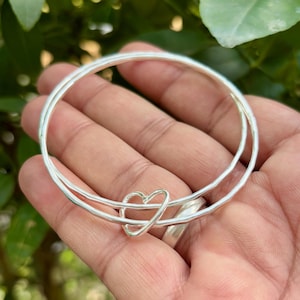 Pode incluir: Uma pulseira de prata com um berloque em forma de coração. A pulseira apresenta duas bandas conectadas e um delicado design de coração no centro. A joia é exibida em um fundo verde natural.