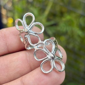 May include: A silver-colored ring featuring a double flower design. Each flower has five openwork petals and a central bead. The ring is shown against a blurred green background.