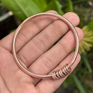 Może przedstawiać: Polerowana miedziana bransoletka typu bangle o gładkim, okrągłym kształcie. Bransoletka zawiera grupę siedmiu małych, połączonych ze sobą miedzianych pierścieni, które przesuwają się wzdłuż głównej obręczy, tworząc prosty i metaliczny wygląd.