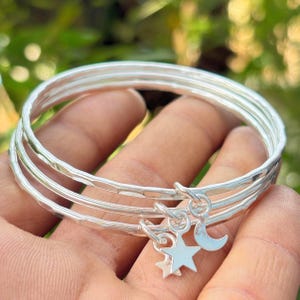 May include: Three silver bangles with a hammered finish. Each bangle features a small charm, including a star and a crescent moon. The bangles are shown against a blurred green backdrop.
