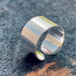 Puede incluir: Un anillo ajustable plateado con un acabado de metal cepillado. El anillo tiene una banda ancha que se curva para crear un diseño abierto. El anillo se muestra sobre una superficie oscura y texturizada.