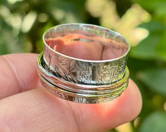 May include: A silver-colored spinner ring with floral and leaf patterns. The ring has a wide band with a textured design and a spinning outer band. The ring is held on a finger against a blurred green background.