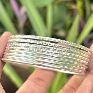 May include: A stack of silver bangles with textured and smooth surfaces. The bangles are stacked together, showcasing a variety of designs. The bracelets are displayed against a blurred green background.