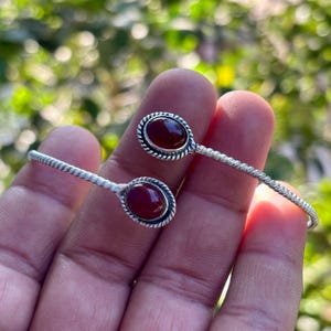 May include: A silver bracelet featuring two oval, dark red gemstones. The gemstones are set in a decorative frame. The bracelet has a twisted design on the band.