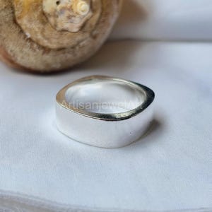 May include: A silver ring with a flat, rectangular band. The ring has a smooth, polished surface and a slightly curved design. The ring is displayed on a white surface.