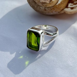 Op de afbeelding: Een zilveren ring met een grote, rechthoekige, heldergroene edelsteen. De ring heeft een eenvoudig, modern ontwerp met een gesplitste band. De edelsteen is gezet in een vierkante bezel setting en de ring wordt gepresenteerd op een wit oppervlak.
