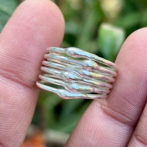 May include: A set of stackable silver rings. The rings have an organic, flowing design with a slightly textured surface. The rings are held between fingers, with a blurred green background.