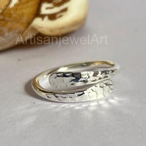 May include: A silver wrap ring with a hammered texture. The ring features a smooth, polished band that encircles the finger. The text "ArtisanjewelArt" is visible in the background.