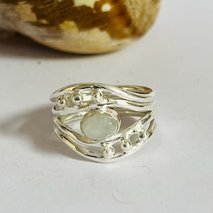 May include: A silver ring with a light green oval gemstone at the center. The ring has a layered design with small silver beads and wavy lines. The ring is on a white surface.