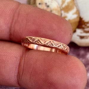 Puede incluir: Primer plano de un anillo de oro rosa con un patrón grabado detallado. La superficie del anillo está adornada con un diseño repetido de hojas estilizadas o formas de plumas. El anillo se muestra en un dedo, mostrando su intrincada artesanía.