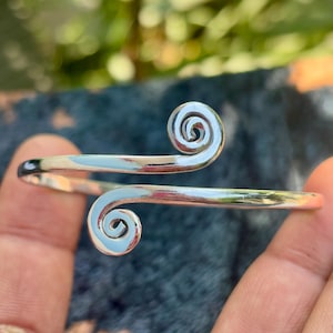 Op de afbeelding: Een zilverkleurige manchetarmband met een spiraalvormig ontwerp aan elk uiteinde. De armband heeft een gladde, gepolijste afwerking en wordt getoond tegen een vage achtergrond. De spiralen zijn verhoogd en gedetailleerd, wat de sieraden visueel aantrekkelijk maakt.