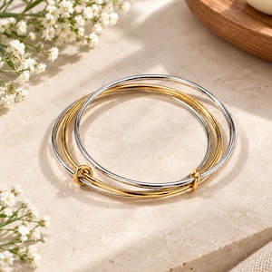 May include: A set of three interlocking bangle bracelets featuring a combination of polished silver-tone and gold-tone metal bands. The circular bracelets are connected by small, fixed metal loops, creating a continuous, layered design.